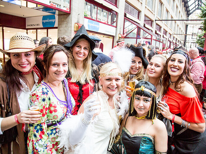 Women in colorful costumes celebrate at a carnival and hold champagne glasses up to the camera.